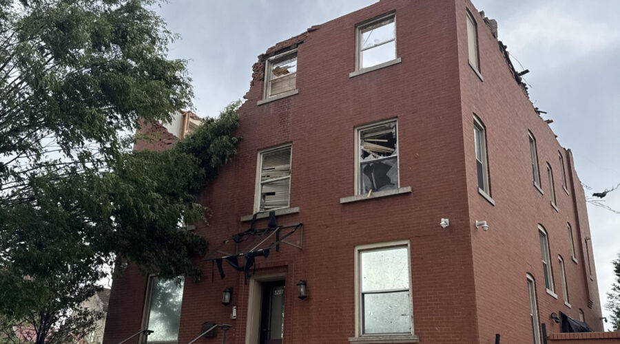 Damaged red brick office building with missing roof and third floor after a tornado, shattered windows, and debris around the entrance.